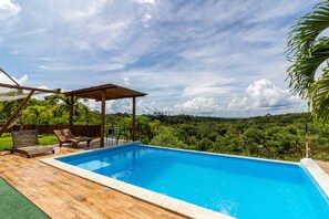 A swimming pool with wooden deck and lounge chairs, surrounded by lush greenery and a clear sky. - PF12 - Ótima Casa 3 Quartos - Açu da Torre (Mata de São João)