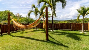 A hammock between trees in a grassy area with a wooden fence and palm trees. - PF12 - Ótima Casa 3 Quartos - Açu da Torre (Mata de São João)