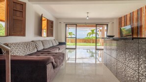 A modern living room with a sofa, a TV, and a view of a tropical garden. - PF12 - Ótima Casa 3 Quartos - Açu da Torre (Mata de São João)