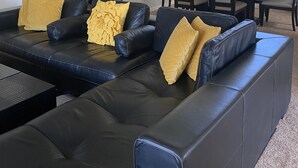 Ein Wohnzimmer mit einem schwarzen Ledersofa, einem Esstisch mit Stühlen, einem Deckenventilator und einer Tür zu einem anderen Raum.