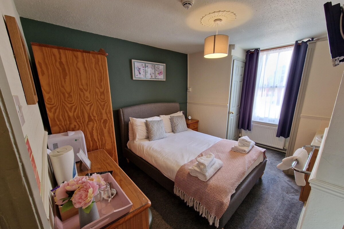 A hotel room with a bed, bedside table, and a desk with a lamp and flowers.