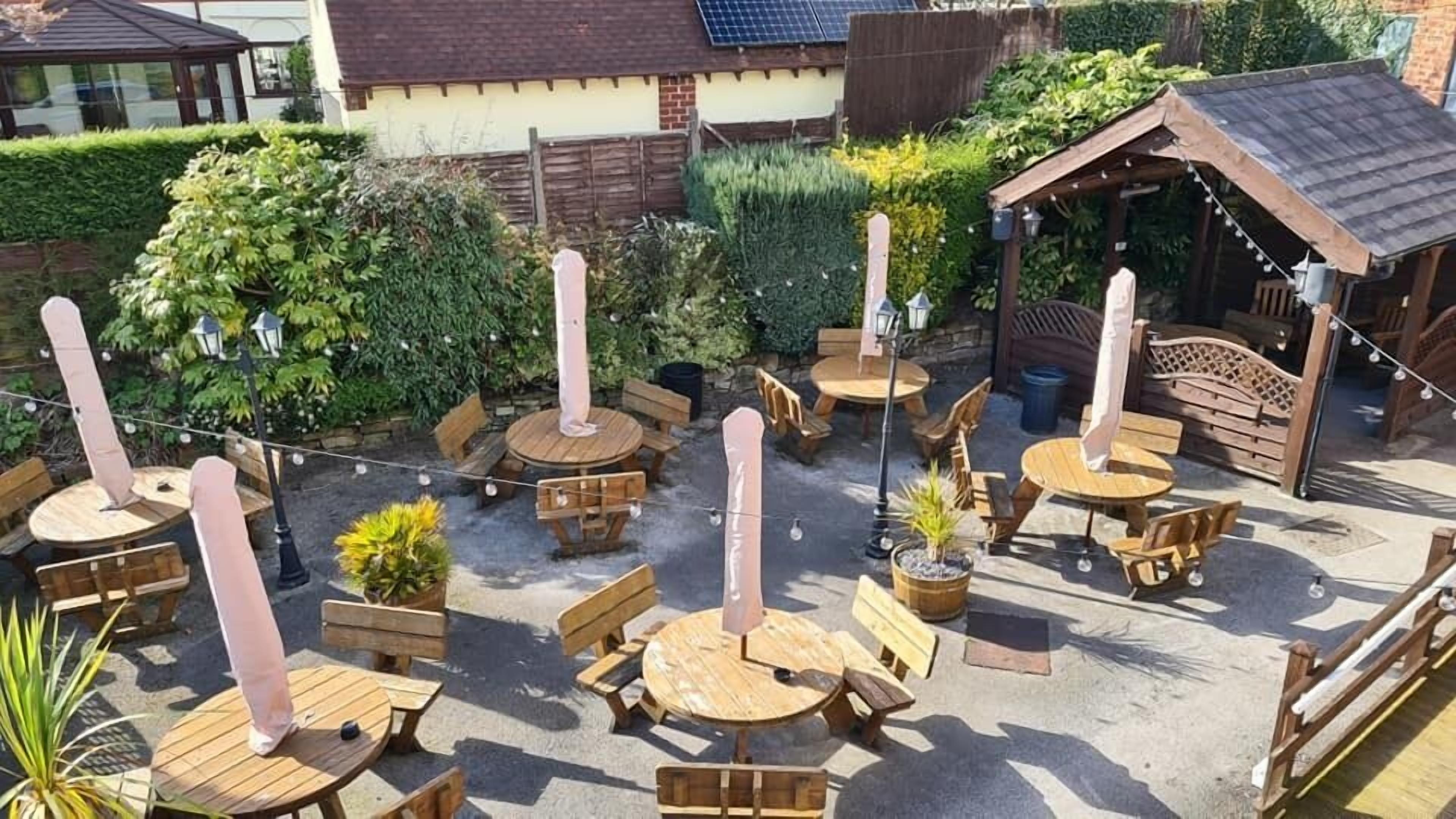 En udendørs patio med flere træborde og bænke, omgivet af grøn vegetation og en træpergola.
