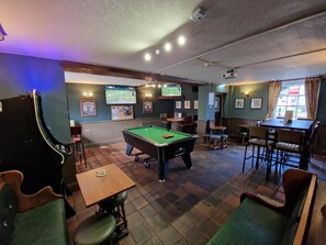 A billiards room with a pool table, chairs, and a television screen displaying a sports game. - The Foresters Arms (Tarporley)