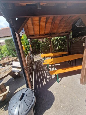 An outdoor patio area with wooden tables and benches, a covered seating area, and a view of surrounding greenery. - The Foresters Arms (Tarporley)