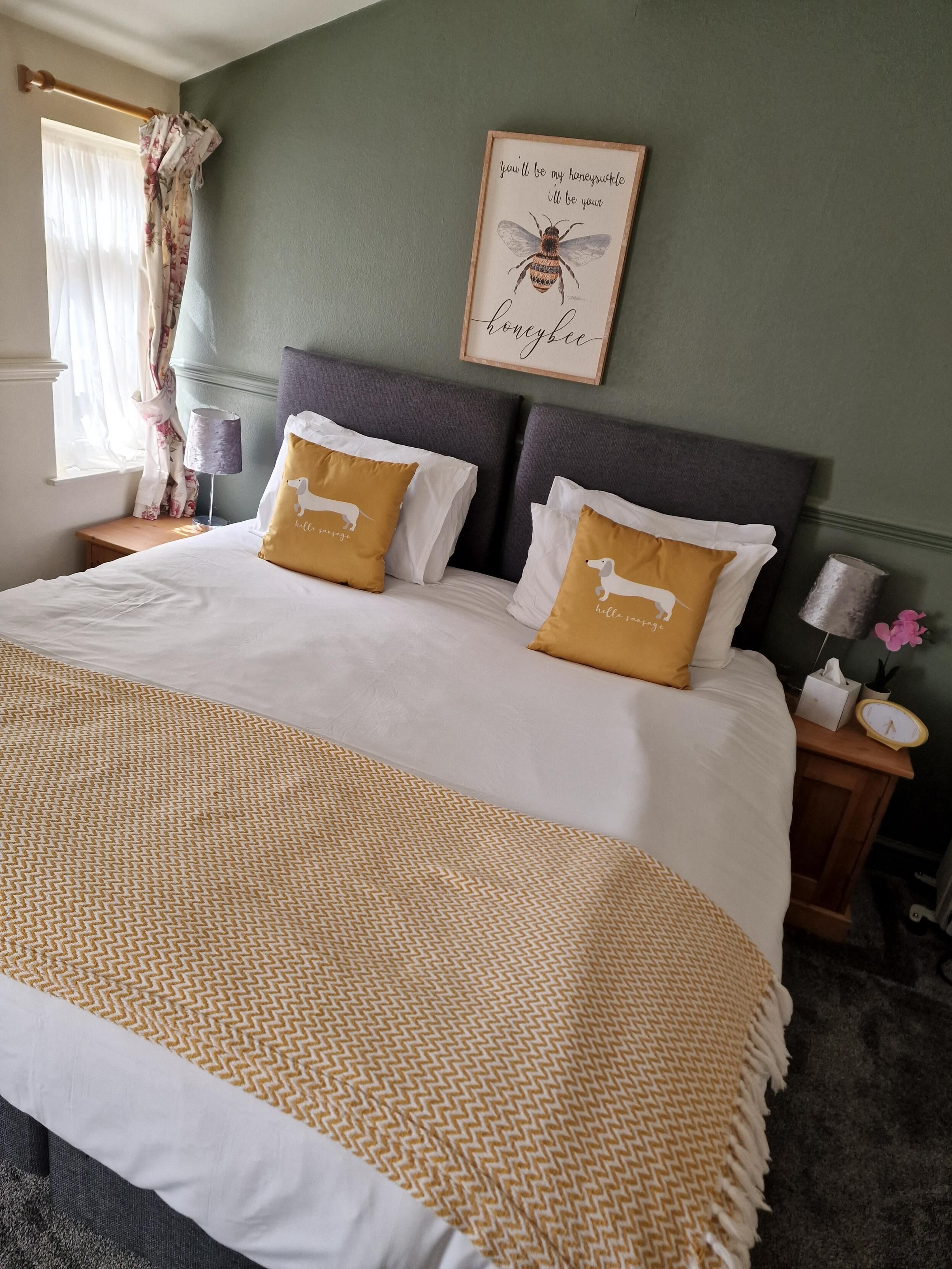 A neatly made bed with white and gold bedding, two decorative pillows, a framed picture on the wall, and a bedside table with a lamp and a clock.
