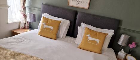 A neatly made bed with white and gold bedding, two decorative pillows, a framed picture on the wall, and a bedside table with a lamp and a clock.