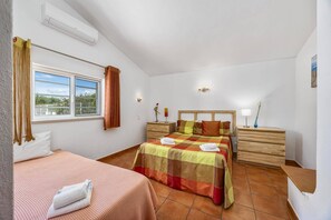 A bedroom with a bed, a bedside table, a window with curtains, and an air conditioning unit. - Villa Dela by Villa Plus (Albufeira)