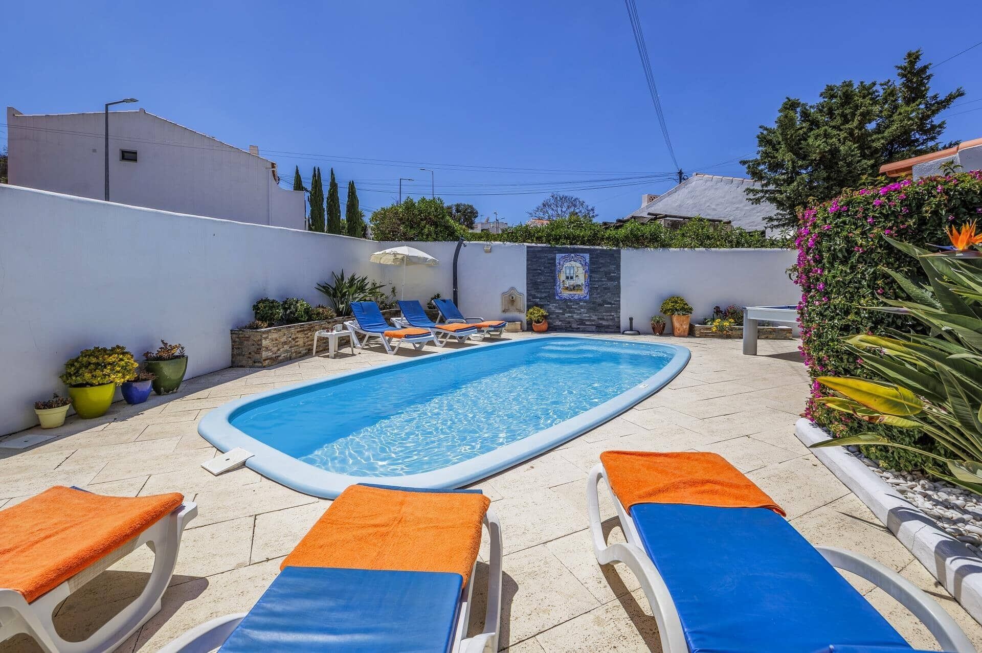 A rectangular swimming pool with blue water, surrounded by white lounge chairs with orange and blue towels.