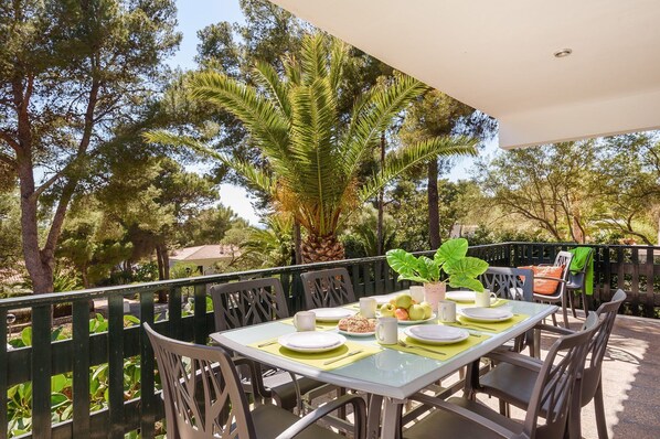 An outdoor dining area with a table set for two, surrounded by chairs, with a view of trees and a fence.
