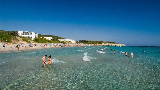 Una playa con agua cristalina, gente nadando y caminando, y edificios al fondo.