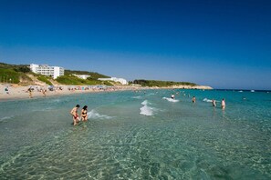 A beach with clear water, people swimming and walking, and buildings in the background.
