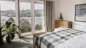 Une chambre d’hôtel avec un lit, une commode en bois, une plante en pot et une vue sur une rivière et des bâtiments, visible à travers de grandes portes coulissantes.