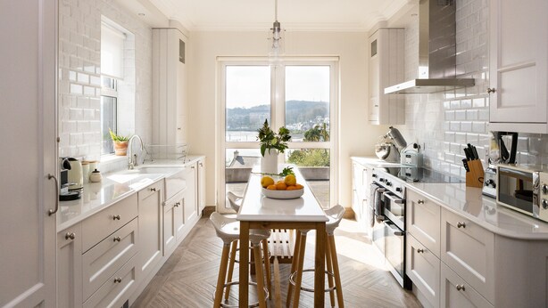 Une cuisine moderne avec des meubles blancs, un îlot central doté de tabourets, une grande fenêtre offrant une vue sur la ville et un luminaire suspendu.