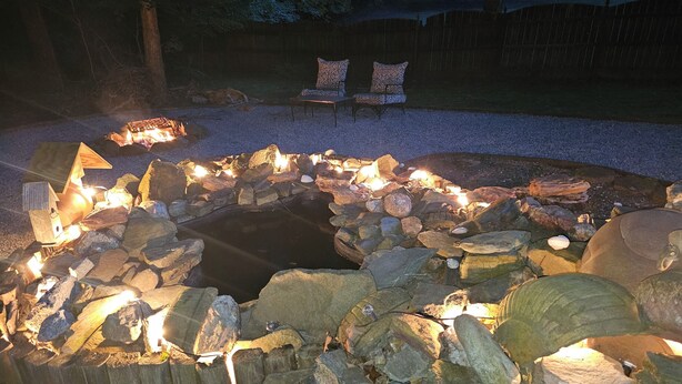 A stone fire pit with logs and a small pond, surrounded by rocks and two chairs with cushions.