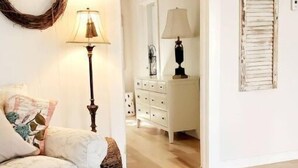 A cozy living room with a sofa, a round table, and a lamp. There is a dresser with drawers and a mirror. A wreath hangs on the wall.