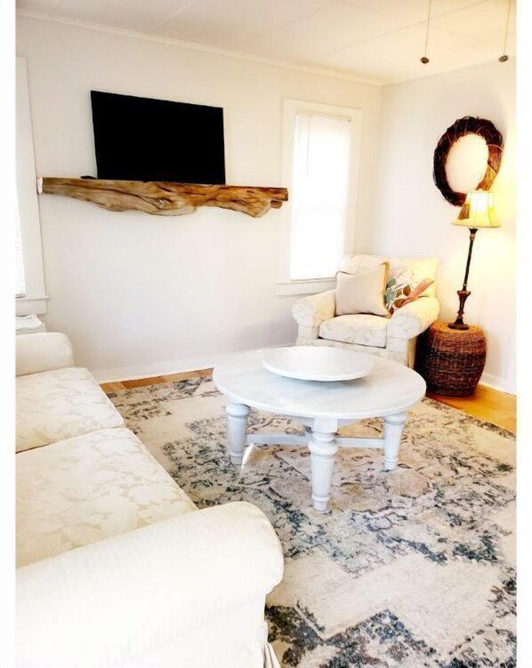 Una sala de estar con un sofá blanco, una mesa de centro redonda, una silla con cojines, un televisor de pantalla plana montado en la pared y una lámpara sobre una mesita auxiliar.