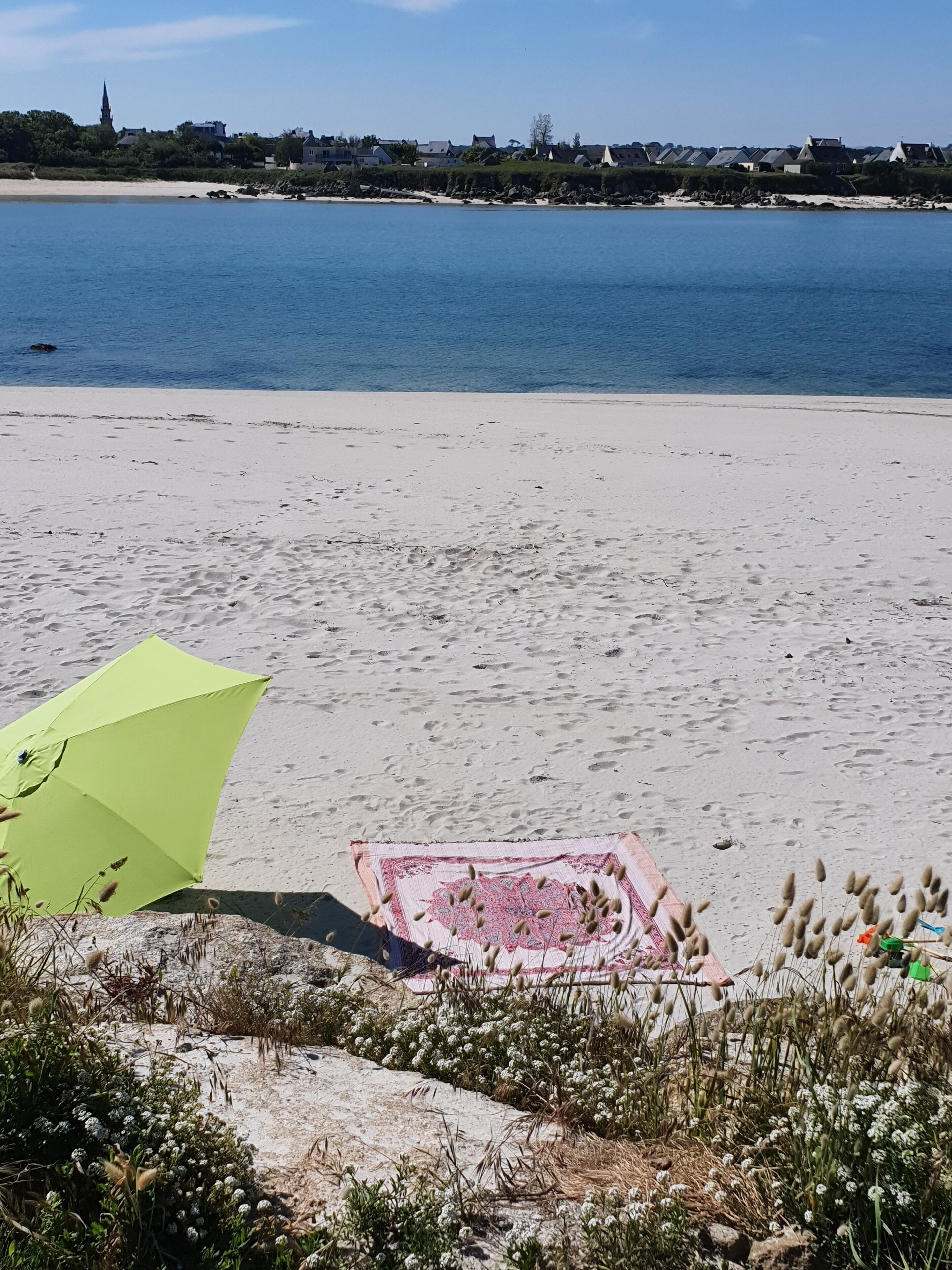 Ein Strand mit einem grünen Sonnenschirm und einer rosa Decke im Sand.