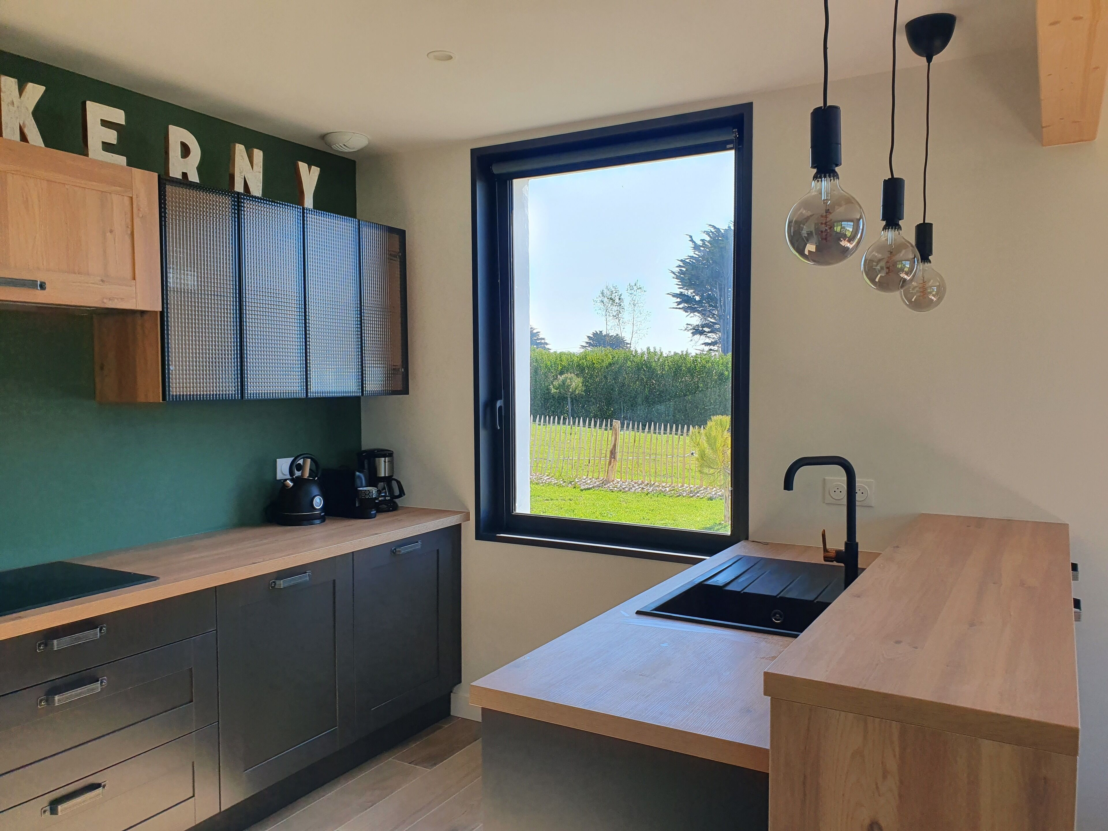 A modern kitchen with a wooden countertop, dark cabinets, a built-in stove, and a window with a view of a garden.