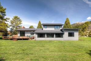 A modern two-story house with a large front lawn, a deck, and a mountain backdrop.