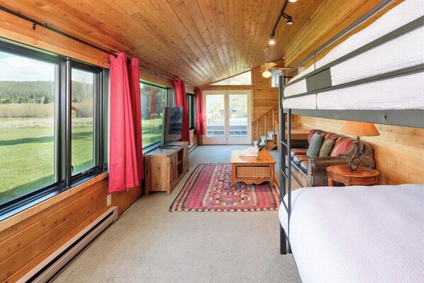 A cozy cabin interior with wooden walls, large windows, a bed, a sofa, and a small table.
