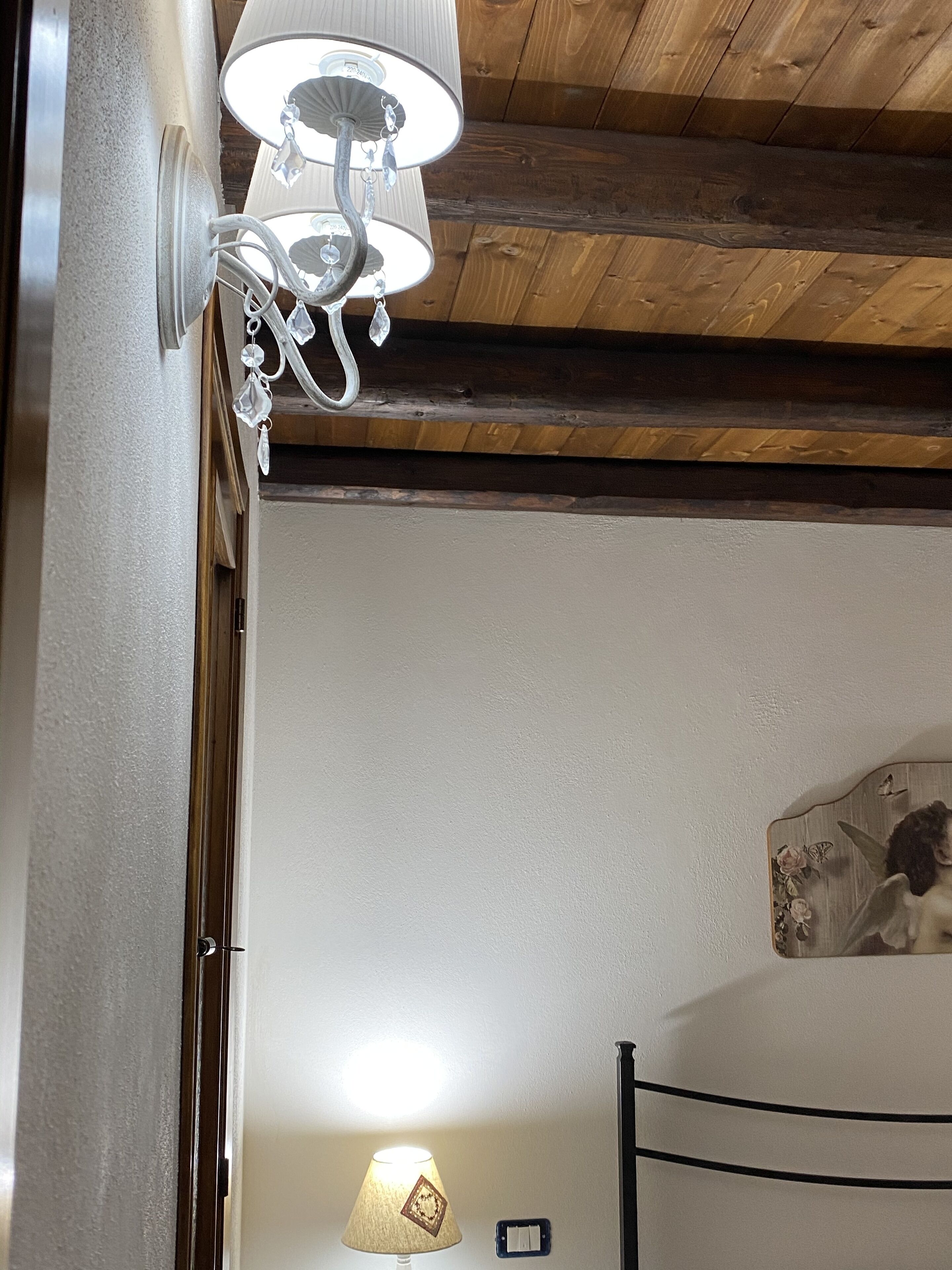 Une pièce avec un plafond en bois, une lampe fixée au mur, un lit avec une tête de lit et un petit cadre photo accroché au mur.