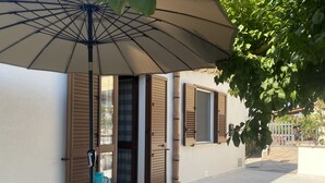 Une terrasse extérieure avec une table dressée pour le repas, un grand parasol et un bâtiment doté de volets en bois.