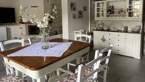 Un coin repas avec une table en bois, des chaises blanches, un vase avec des fleurs, une cheminée et un espace cuisine avec des placards et des étagères.