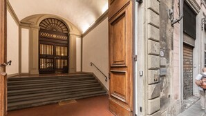 Una entrada de edificio con una puerta de madera, un conjunto de escaleras que conducen a una puerta con un arco decorativo, y un suelo de baldosas.