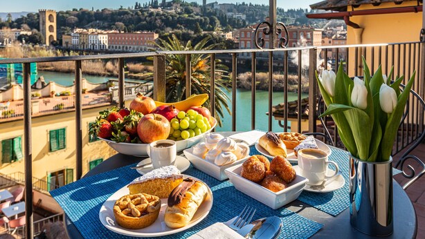 Un balcón con una mesa dispuesta para el desayuno, que incluye bollería, frutas y café, con vistas a un pueblo y un río pintorescos.