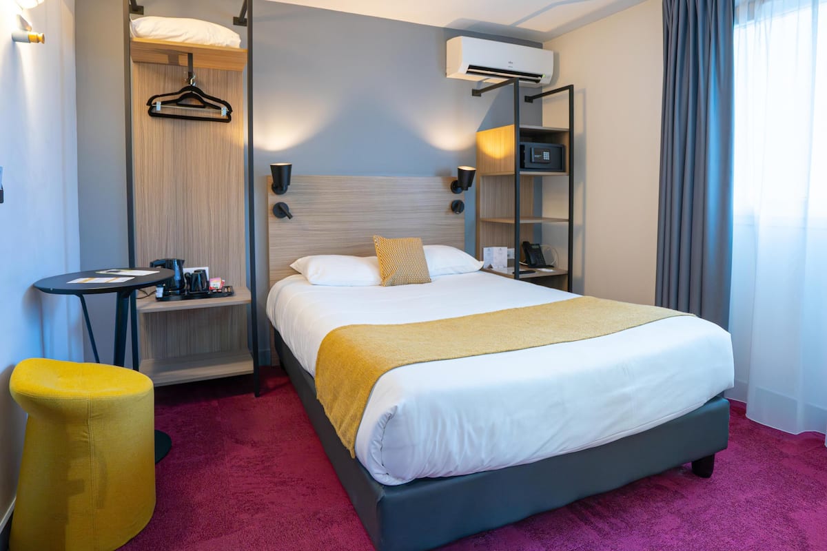 Une chambre d’hôtel équipée d’un lit, d’une table de chevet, d’un porte-vêtements, d’un appareil de climatisation et d’un petit tabouret.