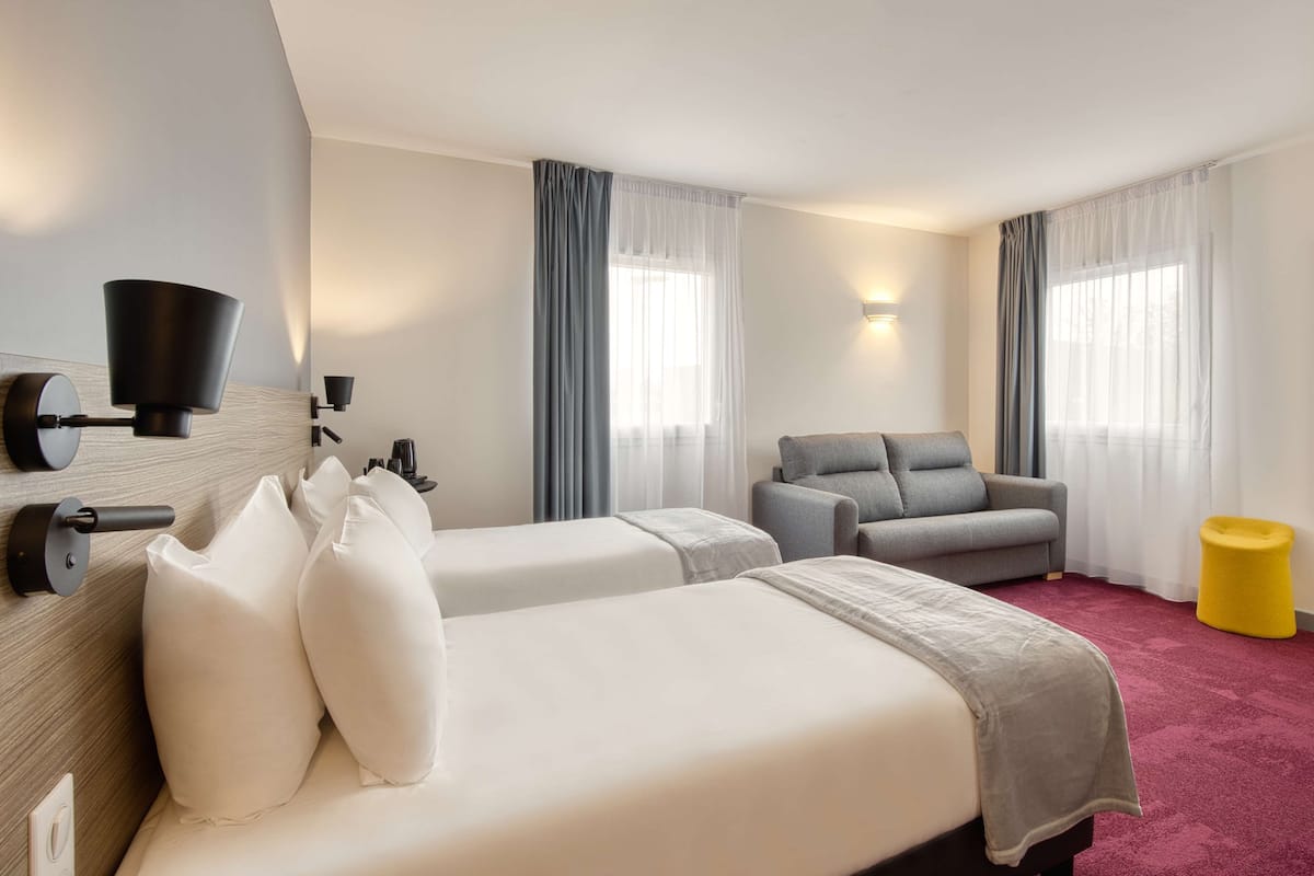 Une chambre d’hôtel avec deux lits, un canapé, un pouf jaune et un téléviseur fixé au mur.