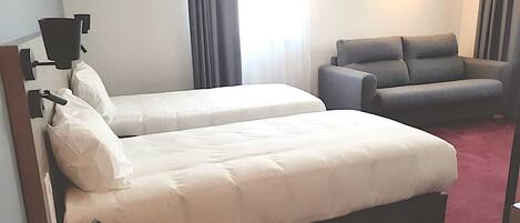 A hotel room with two beds, a grey armchair, a desk with a telephone, and a window with curtains.