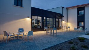 A modern outdoor seating area with chairs and tables, a gravel ground, and a building with large windows and a dark facade. - Sure Hotel by Best Western Les Portes de Montauban (Bressols)