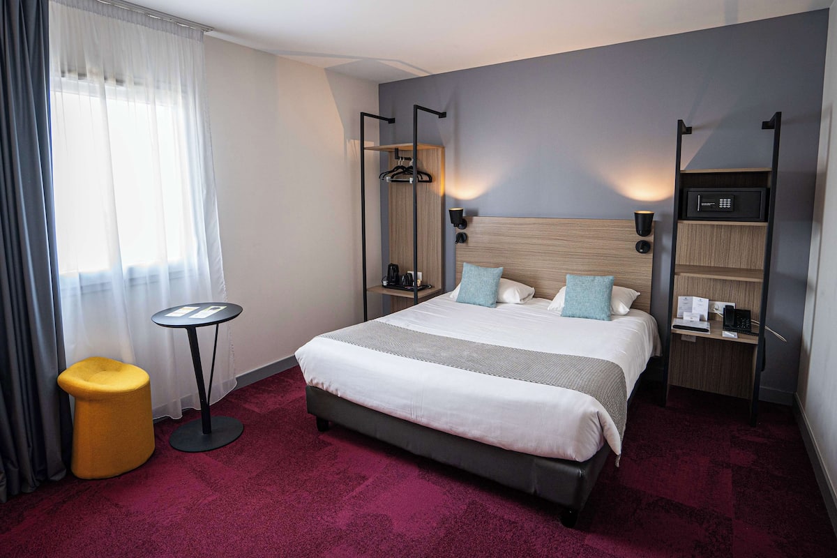 Une chambre d’hôtel avec un grand lit, une petite table ronde, un tabouret jaune, une armoire et une étagère avec une radio.