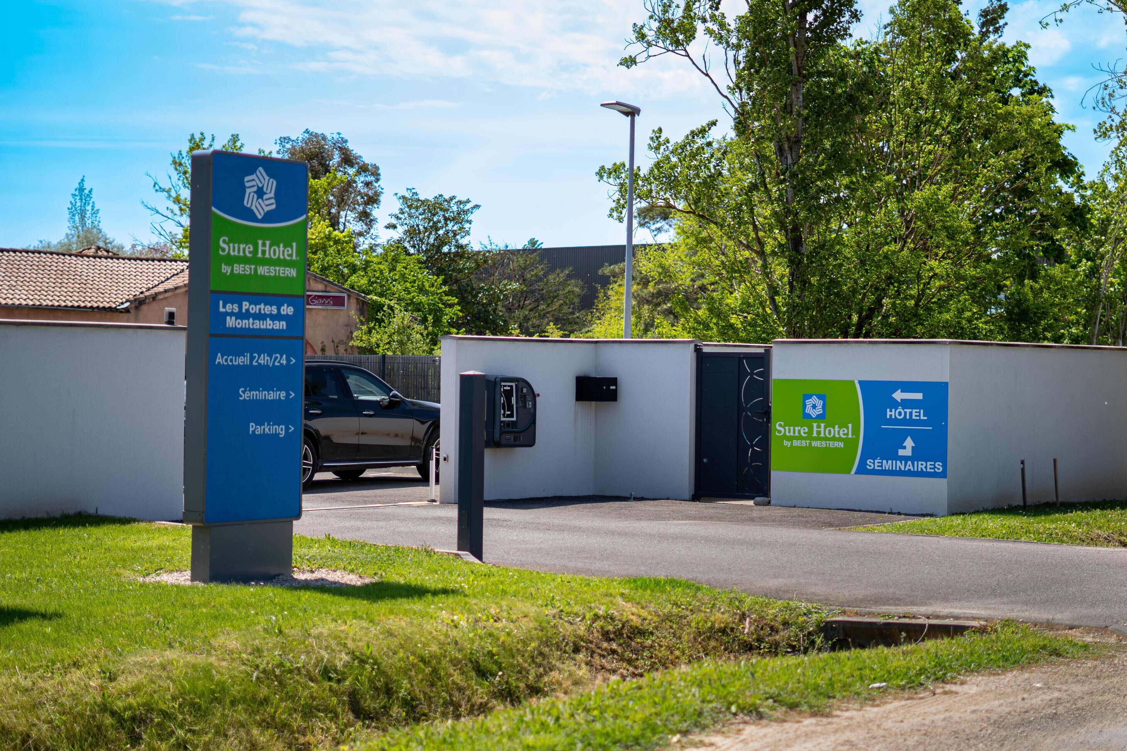 A hotel entrance with a sign indicating 'Sure Hotel by Best Western,' a parking area, and a building entrance.