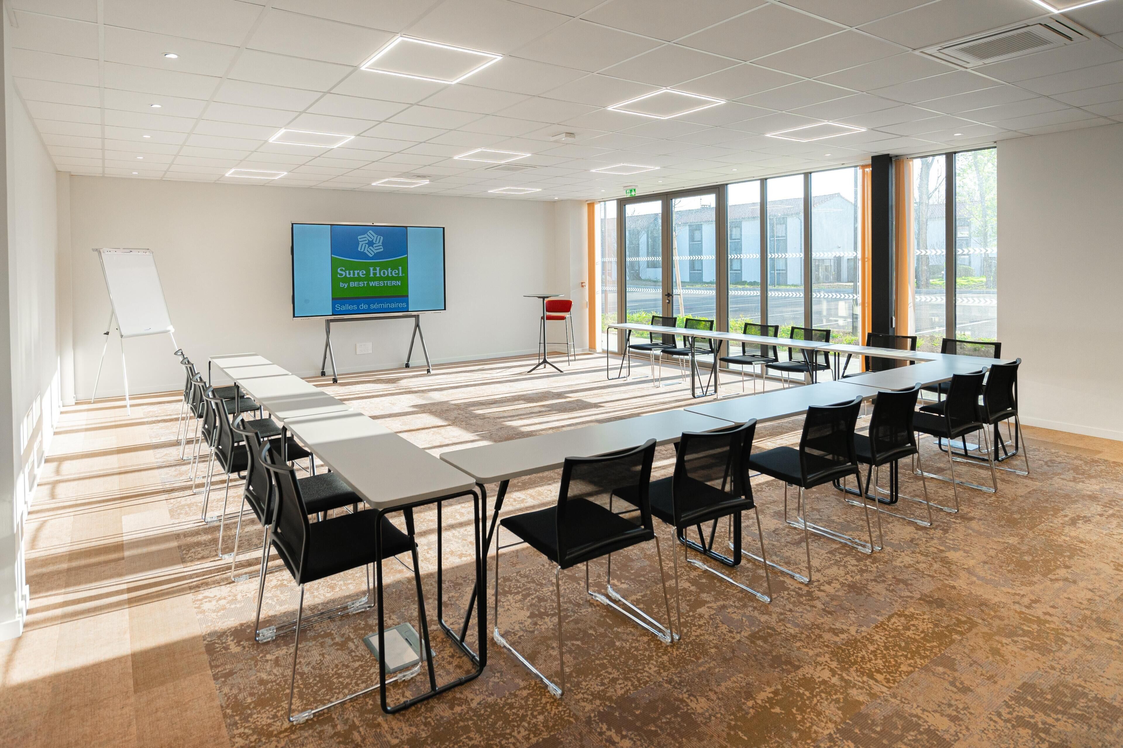 A conference room with a long table, chairs, a whiteboard, and a screen displaying Sure Hotel by Bestest Hotels.