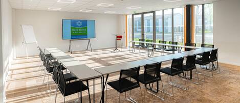 A conference room with a long table, chairs, a whiteboard, and a screen displaying Sure Hotel by Bestest Hotels.