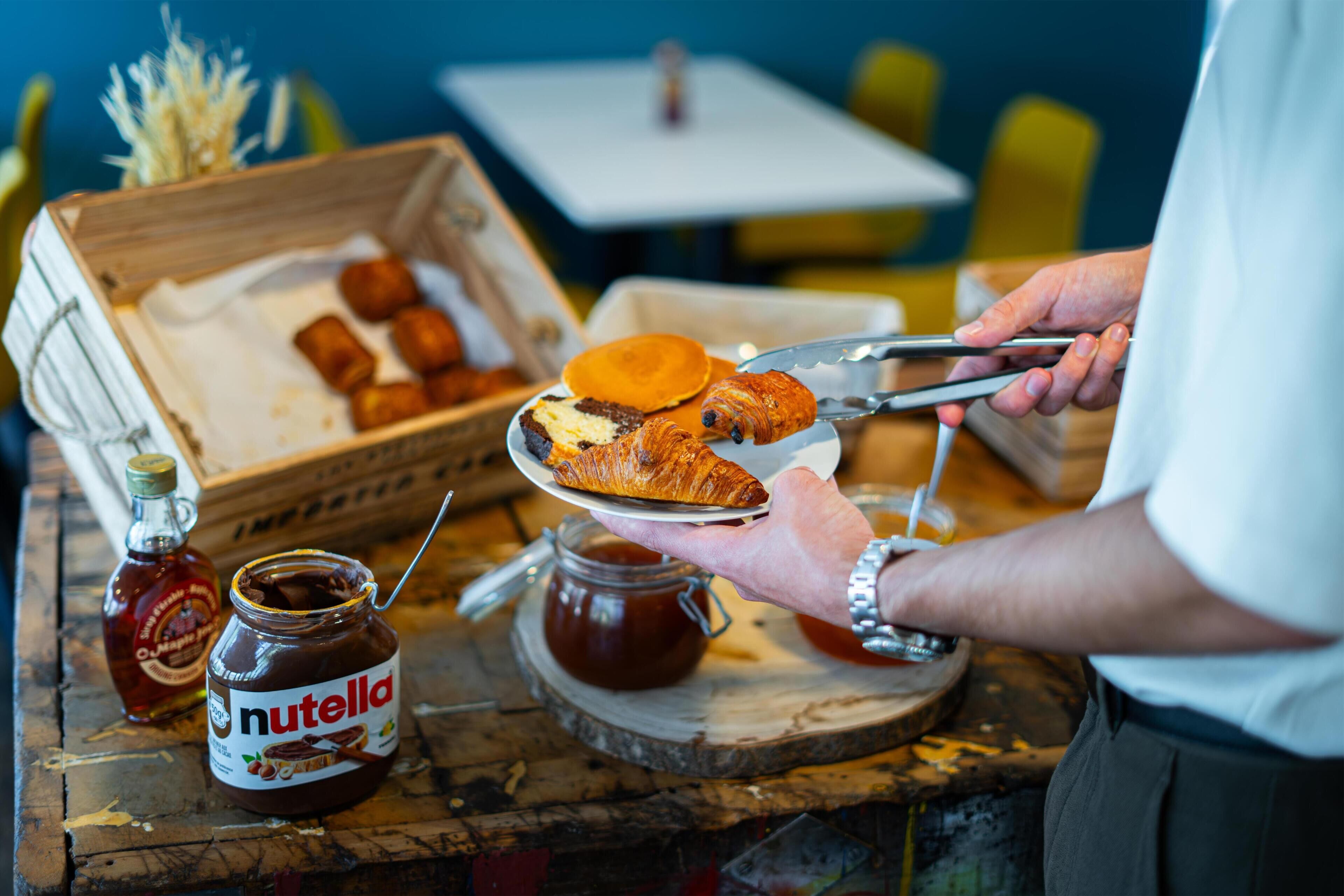 A person holding a plate with a croissant and a piece of cake, a jar of Nutella, and a bottle of syrup on a wooden table.