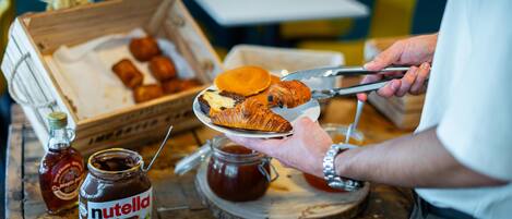 A person holding a plate with a croissant and a piece of cake, a jar of Nutella, and a bottle of syrup on a wooden table.