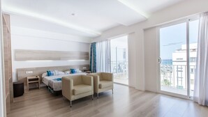 Quarto de hotel moderno com uma cama grande, duas poltronas, uma mesinha de cabeceira e vista para o mar através de amplas janelas.