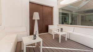 Uma área de estar externa moderna e bem iluminada, com mobiliário branco, incluindo sofás e mesas, e uma grande janela que oferece vista do interior.