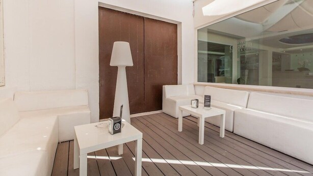 A modern, well-lit outdoor seating area with white furniture, including sofas and tables, and a large window providing a view of the interior. - Gracia (Llucmajor)