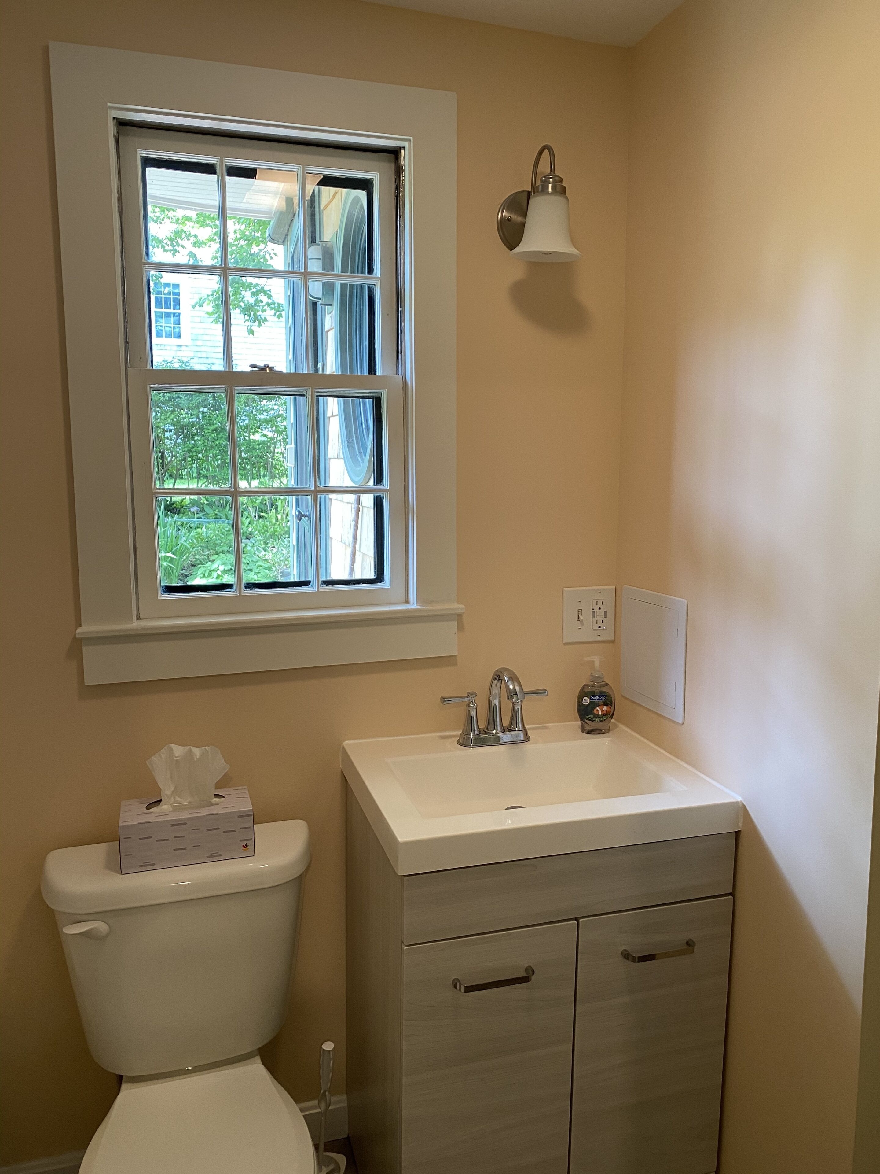 A bathroom with a toilet, sink, cabinet, window, and a wall-mounted light fixture.