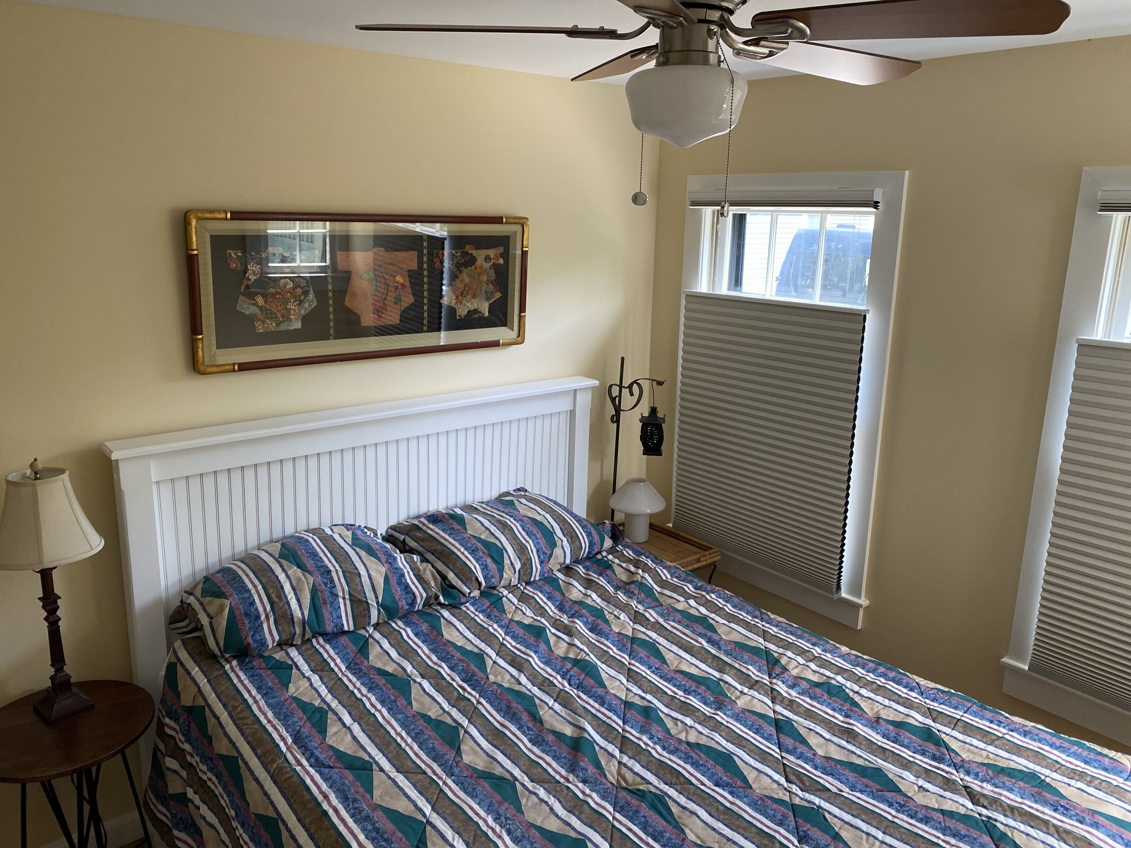 A bedroom with a bed, a ceiling fan, a window with blinds, a wall-mounted picture, and a bedside table with a lamp.