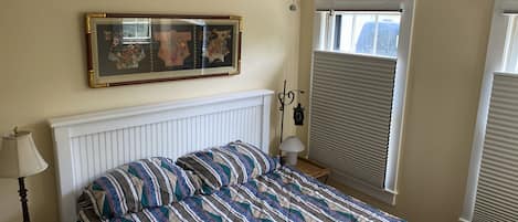 A bedroom with a bed, a ceiling fan, a window with blinds, a wall-mounted picture, and a bedside table with a lamp.