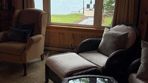 A cozy room with a view of a lake, a wicker sofa, a cushioned chair, and a glass coffee table with magazines.