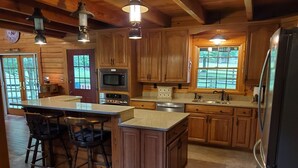 A spacious kitchen with wooden cabinets, a large island with bar stools, a microwave, oven, and a refrigerator. - Spring Acre Log Cabin (Paradise)