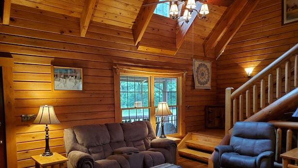 A cozy log cabin interior with a wooden ceiling, a sofa, a recliner, a wooden chest, and a staircase. - Spring Acre Log Cabin (Paradise)