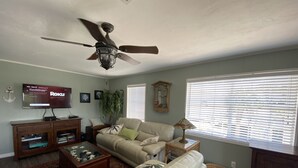 Ein Wohnzimmer mit Deckenventilator, einem Fernseher mit Roku, einer Couch, einem Couchtisch und einem Fenster mit Jalousien.