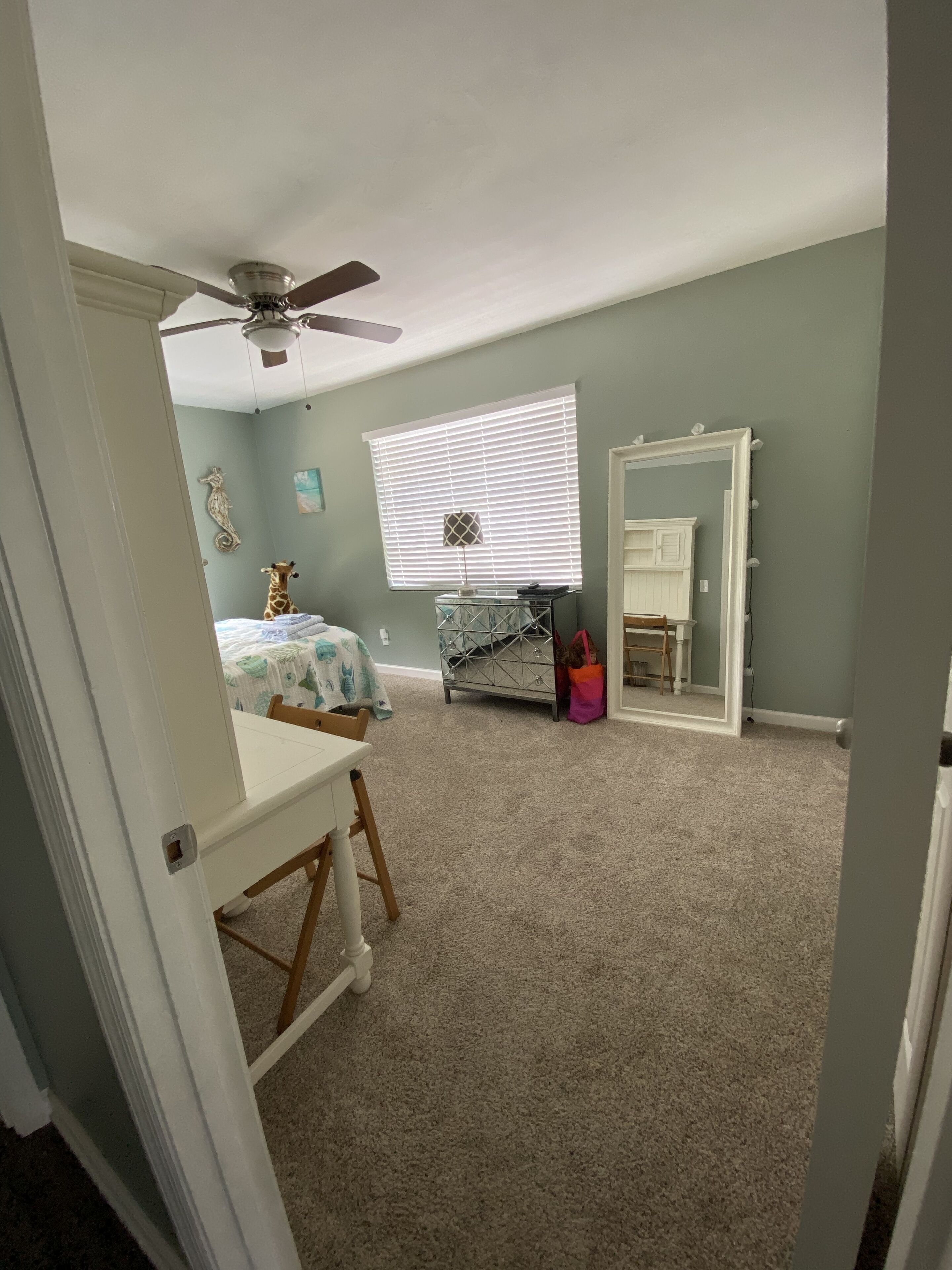 A bedroom with a bed, a desk, a chair, a ceiling fan, a window with blinds, a mirror, and a dresser.